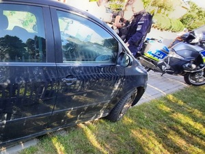 przed granatowym osobowym samochodem stoi policjant. Obok niego stoją kobieta i mężczyzna. Za nim stoi policyjny oznakowany motocykl