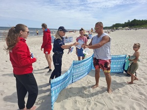 na plaży policjantka w towarzystwie ratownika i ratowniczki WOPR wręcza ulotkę plażowiczowi