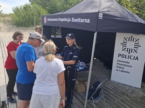 przed wejściem na plażę stoi stoisko sanepidu oraz roll up Komendy Powiatowej Policji w Lęborku. Uśmiechnięta policjantka rozmawia z grupą seniorów