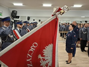 święto Policji obchodzone w auli. Przed pocztem sztandarowym stoi Zastępczyni Komendanta Wojewódzkiego Policji w Gdańsku. Funkcjonariusze z pocztu salutują