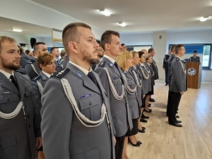 święto Policji obchodzone w auli. Przy ścianie z luster w kilku rzędach stoją mianowani policjanci. Obok znajduje się mównica przy której stoi policjantka