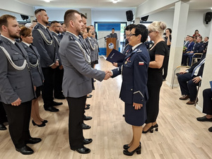 święto Policji obchodzone w auli. Przy ścianie w kilku rzędach stoją mianowani policjanci. Zastępca Komendanta Wojewódzkiego Policji w Gdańsku oraz Komendant Powiatowa Policji w Pucku wręczają im akty mianowania na wyższe stopnie służbowe