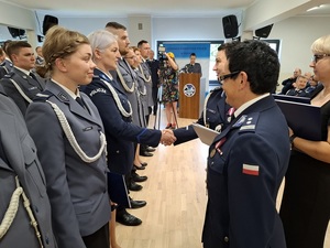 święto Policji obchodzone w auli. Przy ścianie w kilku rzędach stoją mianowani policjanci. Zastępca Komendanta Wojewódzkiego Policji w Gdańsku oraz Komendant Powiatowa Policji w Pucku wręczają im akty mianowania na wyższe stopnie służbowe