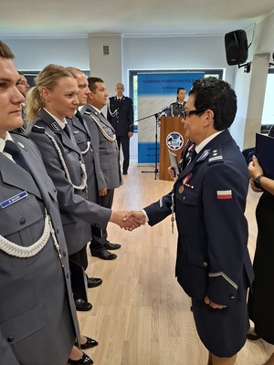 święto Policji obchodzone w auli. Przy ścianie w kilku rzędach stoją mianowani policjanci. Zastępca Komendanta Wojewódzkiego Policji w Gdańsku oraz Komendant Powiatowa Policji w Pucku wręczają im akty mianowania na wyższe stopnie służbowe