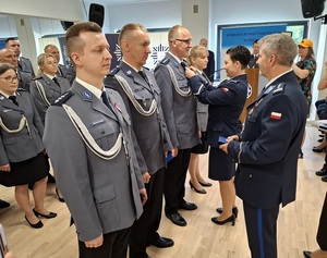 święto Policji obchodzone w auli. Przy ścianie w kilku rzędach stoją mianowani policjanci. Zastępca Komendanta Wojewódzkiego Policji w Gdańsku oraz Komendant Powiatowy Policji w Lęborku wręczają im akty mianowania na wyższe stopnie służbowe