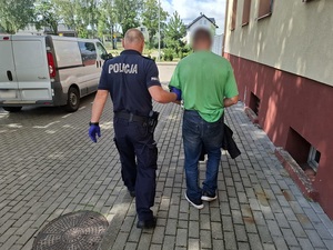 policjant prowadzi mężczyznę z rękoma skutymi kajdankami w stronę nieoznakowanego radiowozu