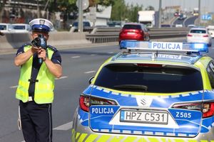 przy oznakowanym radiowozie stoi policjant ruchu drogowego, który mierzy prędkość przejeżdżających pojazdów za pomocą ręcznego miernika prędkości