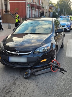 miejsce zdarzenia drogowego. Przy przejściu dla pieszych przed maską czarnego opla astry leży hulajnoga elektryczna. Za pojazdami stoi policyjny radiowóz i dwoje policjantów