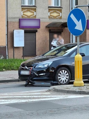 miejsce zdarzenia drogowego. Przy przejściu dla pieszych przed maską czarnego opla astry leży hulajnoga elektryczna