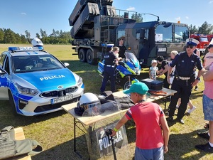 piknik pod chmurką. Przed stołem ze sprzętem policyjnym stoi radiowóz i policyjny motocykl. Policjant rozmawia z ludźmi, policjantka asekuruje dziecko siedzące na motocyklu