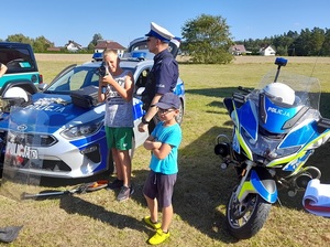 piknik po chmurką przy policyjnym motocyklu i radiowozie chłopiec trzyma ręczny miernik prędkości. Obok stoi policjant ruchu drogowego