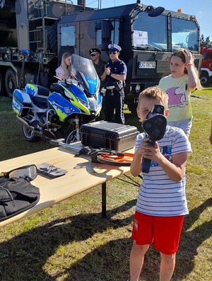 przy policyjnym stoisku profilaktycznym chłopiec trzyma ręczny miernik prędkości. W tle widać policyjny motocykl i radiowóz oraz policjanta ruchu drogowego