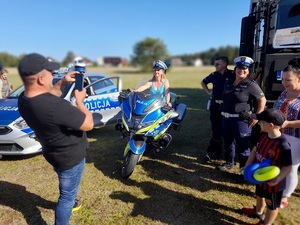 kobieta siedząca na policyjnym motocyklu. Mężczyzna robi jej zdjęcie telefonem. Obok stoją uśmiechnięci policjant i policjantka ruchu drogowego