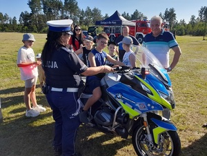 na policyjnym motocyklu siedzi chłopiec. Asekuruje go policjantka ruchu drogowego. Naprzeciw niej stoi uśmiechnięty wójt gm. Wicko oraz inni uczestnicy pikniku