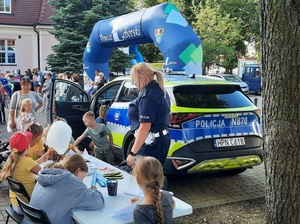 przed budynkiem starostwa przy radiowozie stoi policjantka ruchu drogowego. Do radiowozu wchodzi dziecko. Przed policjantką przy stoliku z kolorowankami i odblaskami siedzą dzieci