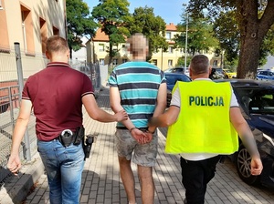 dwaj nieumundurowani policjanci prowadzą mężczyznę z rękoma skutymi z tyłu kajdankami w stronę radiowozu