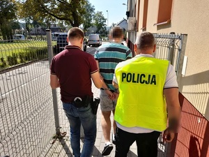 dwaj nieumundurowani policjanci wyprowadzają z budynku komendy mężczyznę z rękoma skutymi z tyłu kajdankami