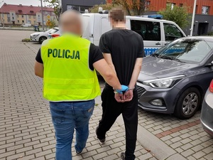 nieumundurowany policjant w kamizelce odblaskowej z napisem policja prowadzi w kierunku radiowozu mężczyznę, który ma ręce skute za plecami kajdankami