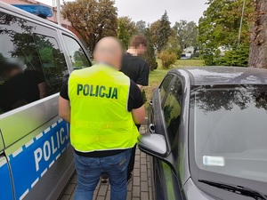 nieumundurowany policjant w kamizelce odblaskowej z napisem policja prowadzi w kierunku radiowozu mężczyznę, który ma ręce skute za plecami kajdankami