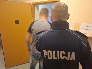 przed otwartymi drzwiami celi stoi zatrzymany mężczyzna. Za nim znajduje się policjant służby dyżurnej