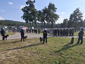 przed głównym wejściem na stadion stoją policjanci w kamizelkach przeciwuderzeniowych i hełmach. Za nimi znajdują się czterej przewodnicy z psami służbowymi. Przy ogrodzeniu od zewnętrznej strony stadionu znajdują się pseudokibice.