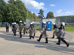 wzdłuż ogrodzenia stadionu idą w szeregu policjanci ubrani w kamizelki przeciwuderzeniowe, hełmy. Trzymają tarcze