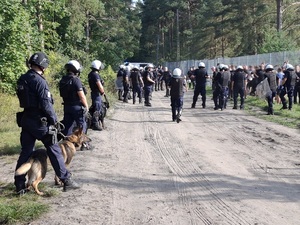 przy ogrodzeniu stadionu od strony sektora kibiców przyjezdnych stoją pseudokibice. Część z nich opiera się rękoma o ogrodzenie. Wzdłuż drogi stoją policjanci z psami służbowymi. Przy pseudokibicach stoją policjanci ubrani w kamizelki przeciwuderzeniowe, hełmy, z tarczami