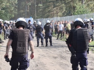 przy ogrodzeniu stadionu od strony sektora kibiców przyjezdnych stoją pseudokibice. Część z nich opiera się rękoma o ogrodzenie. Przy pseudokibicach stoją policjanci ubrani w kamizelki przeciwuderzeniowe, hełmy, z tarczami