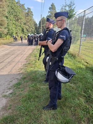 przy ogrodzeniu stadionu stoją dwaj policjanci trzymający długą broń gładkolufową. W tle widać policjantów ubranych w kamizelki przeciwuderzeniowe i kaski