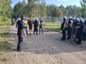 policjant w kamizelce odblaskowej i policjant ubrany w kamizelkę przeciwuderzeniową oraz kask prowadzą mężczyznę. Obok stoją policjanci biorący udział w zabezpieczeniu meczu