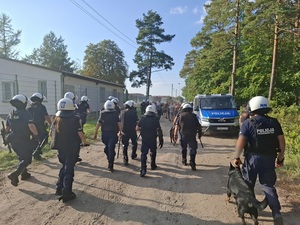 wzdłuż ogrodzenia stadionu od strony lasu idzie grupa pseudokibiców. Obok nich i za nimi idą policjanci zabezpieczający mecz, w tym przewodnik z psem służbowym