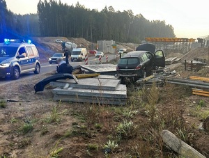 poza jezdnią, na gruntowej nawierzchni, wśród materiałów i sprzętu budowlanego znajduje się rozbity czarny opel astra. Na jezdni stoi policyjny radiowóz