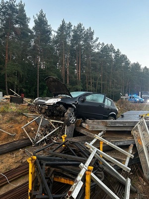 poza jezdnią, na gruntowej nawierzchni, wśród materiałów i sprzętu budowlanego znajduje się rozbity czarny opel astra. Na jezdni stoi policyjny radiowóz z włączonymi sygnałami błyskowymi