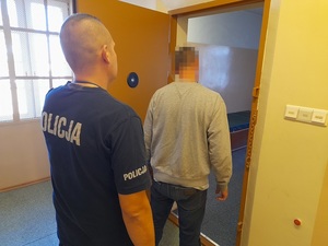 przed otwartymi drzwiami celi stoi mężczyzna w szarej bluzie. Za nim stoi policjant służby dyżurnej