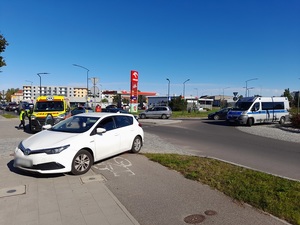 miejsce zdarzenia drogowego. Na chodniku i drodze dla rowerów stoi biała toyota. Za nią znajduje się karetka pogotowia przy której stoi dwoje policjantów ruchu drogowego. Na rondzie stoi policyjny radiowóz