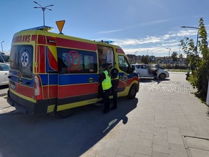 na chodniku stoi karetka pogotowia przy której znajduje się dwoje policjantów ruchu drogowego. Przed karetką na chodniku stoi biała toyota