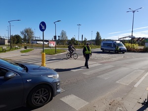 na przejście dla pieszych wchodzi policjant ruchu drogowego który spogląda na zbliżające się auto. Za nim na rondzie stoi radiowóz przy którym znajduje się policjantka