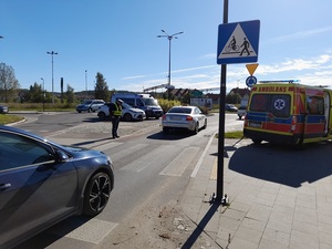 policjant ruchu drogowego spoglądający na przejście dla pieszych. Po przeciwnej stronie stoi karetka pogotowia. Na rondzie stoi radiowóz