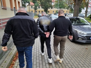 dwaj policjanci kryminalni prowadzą mężczyznę skutego kajdankami w kierunku radiowozu