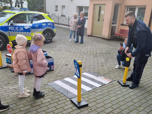 przy zabawkowym przejściu dla pieszych stoi policjant. Naprzeciw stoją dwie dziewczynki