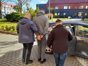 dwaj nieumundurowani policjanci wprowadzają do nieoznakowanego radiowozu mężczyznę skutego kajdankami