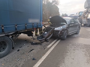 miejsce wypadku drogowego. Na jezdni za przyczepą ciężarówki stoi czarne porsche z rozbitym przodem. Przy aucie znajdują się strażacy. Za autem jedzie koparka