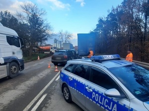 miejsce wypadku drogowego. Na jezdni za przyczepą ciężarówki stoi czarne porsche. Z porsche znajduje się policyjny radiowóz. Przy autach stoją dwaj mężczyźni w odblaskowych pomarańczowych kamizelkach. Drogą jedzie pojazd ciężarowy