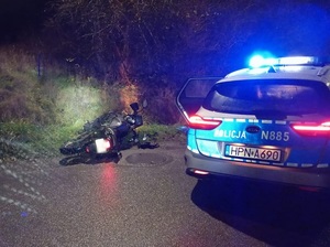 pora nocna. Na poboczu leży przewrócony motocykl. Obok stoi policyjny radiowóz z włączonymi sygnałami błyskowymi