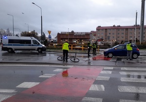 miejsce wypadku drogowego. Dwuetapowe przejście przez pasy. Na pasach przed przejazdem rowerowym stoi niebieski osobowy hyundai. Na chodniku dwuetapowego przejścia dla pieszych i przejazdu dla rowerów leży rower. Na jezdni stoi policyjny radiowóz. Policjanci zabezpieczają miejsce zdarzenia i sporządzają oględziny