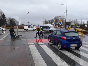 miejsce wypadku drogowego. Dwuetapowe przejście przez pasy. Na pasach przed przejazdem rowerowym stoi niebieski osobowy hyundai. Na chodniku dwuetapowego przejścia dla pieszych i przejazdu dla rowerów leży rower. Na jezdni stoi policyjny radiowóz. Przez pasy przechodzą dwaj policjanci ruchu drogowego. Policjanci z grupy operacyjno - dochodzeniowej prowadzą oględziny roweru