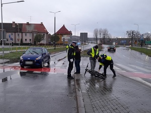 miejsce wypadku drogowego. Dwuetapowe przejście przez pasy. Na pasach przed przejazdem rowerowym stoi niebieski osobowy hyundai. Na chodniku dwuetapowego przejścia dla pieszych i przejazdu dla rowerów leży rower. Na jezdni stoi policyjny radiowóz. Policjanci prowadzą oględziny roweru