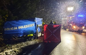 miejsce wypadku drogowego. Na poboczu i jezdni są rozstawione parawany policji i straży pożarnej. Dalej stoi wóz straży pożarnej. Przy parawanach znajdują się strażacy