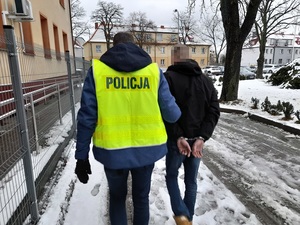 nieumundurowany policjant w kamizelce odblaskowej z napisem policja prowadzi w kierunku radiowozu mężczyznę, który ma ręce skute za plecami kajdankami