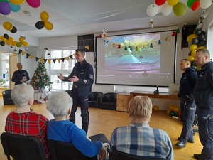 w sali przed grupą seniorów stoi policjant ruchu drogowego. Za nim na ekranie wyświetla się prezentacja